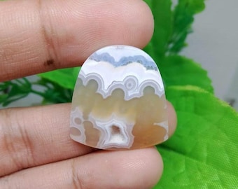 May include: A polished, cabochon-cut gemstone with a unique pattern. The stone features layers of white, light blue, and yellow-brown hues, creating a swirling, cloud-like design. The stone is held in a hand, showcasing its natural beauty.