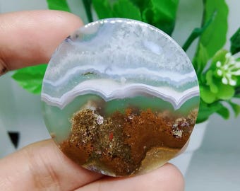May include: A round, green, white, and brown agate cabochon with a layered pattern. The agate has a smooth, polished surface.