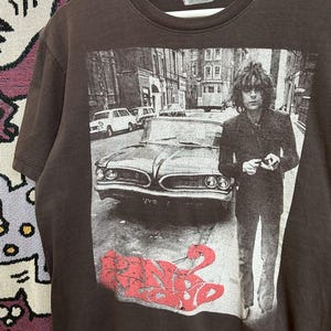 Puede incluir: Camiseta marrón oscuro con una imagen en blanco y negro de una persona y un coche clásico en una calle. La palabra "Syd" está impresa en rojo en la parte inferior de la imagen.