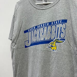 Peut inclure: T-shirt gris avec un graphique bleu et jaune d'un lapin et le texte "South Dakota State Jackrabbits".