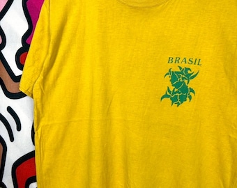 Puede incluir: Una camiseta amarilla de manga corta con cuello redondo y una percha blanca. En la parte superior derecha del pecho, la palabra BRASIL está impresa en verde sobre un diseño gráfico verde estilizado. Una pequeña etiqueta blanca es visible.