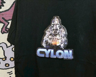 May include: Black t-shirt featuring a graphic of a robotic figure with the word "CYLON" in a blue and silver font. The image is centered on the front of the shirt. The shirt has a crew neck and short sleeves.