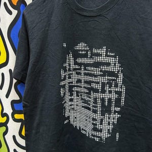 May include: Black crew neck t-shirt with a white abstract design on the front. The design appears to be made up of small, rectangular shapes. The tag on the back of the neck reads "ROY LICHTENSTEIN".