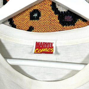 May include: Close-up of a white t-shirt collar with a Marvel Comics tag. The tag is rectangular with red, yellow, and white text. The background is a patterned fabric.