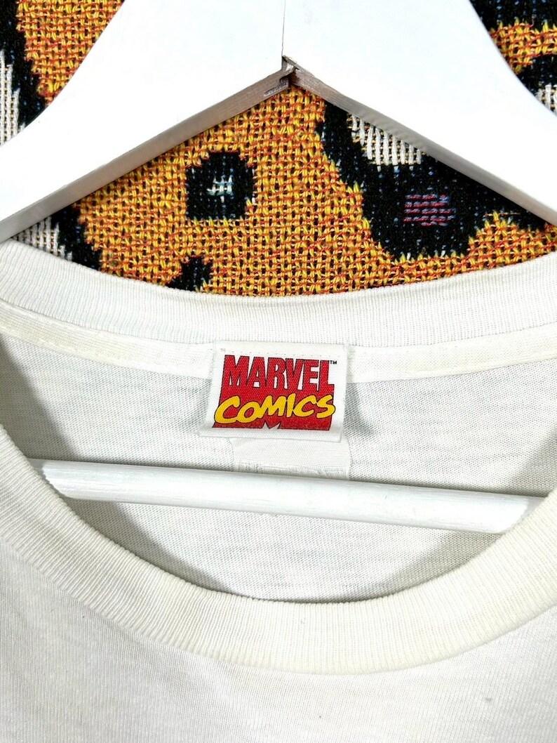May include: Close-up of a white t-shirt collar with a Marvel Comics tag. The tag is rectangular with red, yellow, and white text. The background is a patterned fabric.