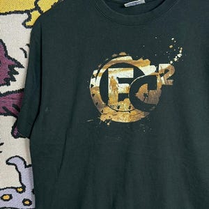 May include: Black crew neck t-shirt with a gold and brown graphic. The graphic features the letters "FC" with a silhouette of a person and the number "2". The design has a distressed look with paint splatter.