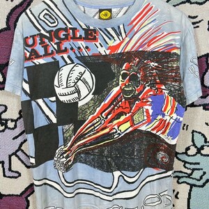 Puede incluir: Camiseta azul claro con un gr&aacute;fico colorido de un jugador de voleibol en acci&oacute;n. La camiseta muestra las palabras "JUNGLE BALL" en rojo y blanco. El dise&ntilde;o incluye un bal&oacute;n de voleibol y un patr&oacute;n de cuadros blancos y negros.