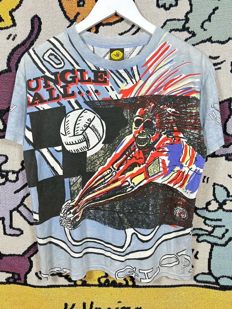 Puede incluir: Camiseta azul claro con un gr&aacute;fico colorido de un jugador de voleibol en acci&oacute;n. La camiseta muestra las palabras "JUNGLE BALL" en rojo y blanco. El dise&ntilde;o incluye un bal&oacute;n de voleibol y un patr&oacute;n de cuadros blancos y negros.