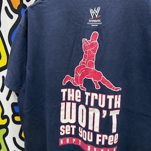 Puede incluir: Camiseta azul marino con un gráfico rojo de dos luchadores. El texto "The Truth Won't Set You Free" está impreso en blanco y rojo, con "Kurt Angle" debajo. El logotipo de WWE Authentic está en la parte superior.