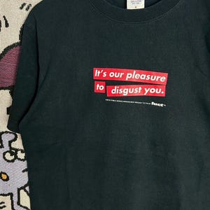 May include: Black t-shirt with the text "It's our pleasure to disgust you" in red block letters. The shirt also has the text "THE PUBLIC SERVICE ANNOUNCEMENT BROUGHT TO YOU BY fuct." The shirt is hanging on a white hanger.