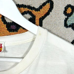 May include: Close-up of a white t-shirt hanging on a white hanger. The t-shirt has a crew neck and appears to be made of a soft, lightweight fabric. The background is a patterned textile with black, orange, and blue elements.