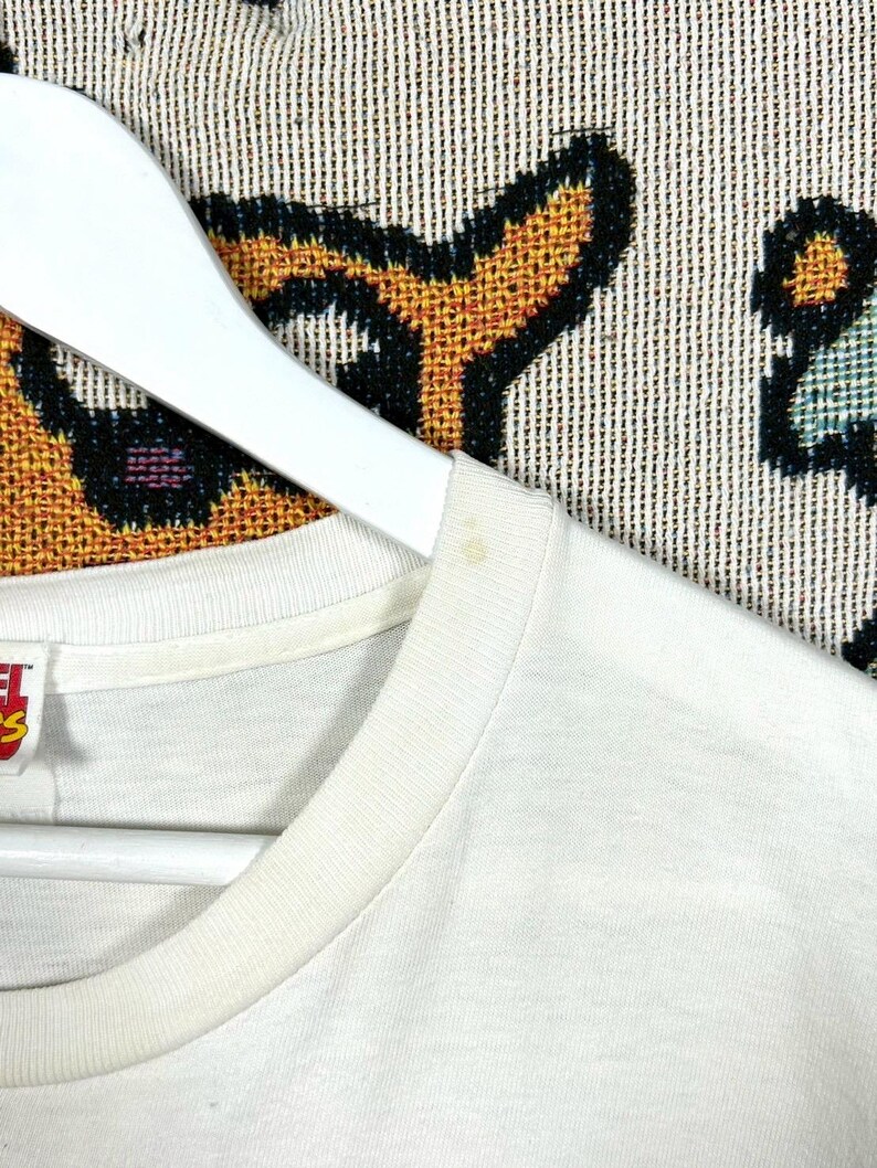 May include: Close-up of a white t-shirt hanging on a white hanger. The t-shirt has a crew neck and appears to be made of a soft, lightweight fabric. The background is a patterned textile with black, orange, and blue elements.