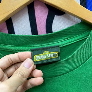 May include: A green t-shirt with a tag that reads "Sesame Street" in white and yellow lettering. The tag is rectangular with a dark green border and the number "123" above the text. The shirt is hanging on a wooden hanger.
