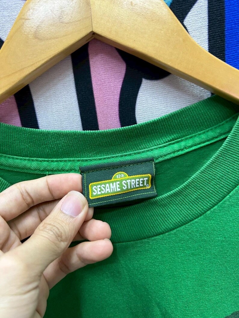 May include: A green t-shirt with a tag that reads "Sesame Street" in white and yellow lettering. The tag is rectangular with a dark green border and the number "123" above the text. The shirt is hanging on a wooden hanger.