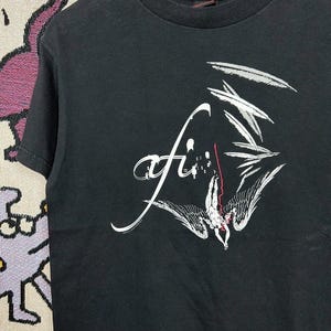 May include: Black crew neck t-shirt with a white graphic design. The design features the word "afi" in a stylized script, along with an illustration of a figure with wings. The figure is in a falling position, with red accents.