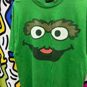 May include: Green Sesame Street t-shirt featuring a large graphic of Oscar the Grouch's face. The design includes white eyes, brown eyebrows, and a black mouth with a red tongue. The shirt has a Sesame Street tag.