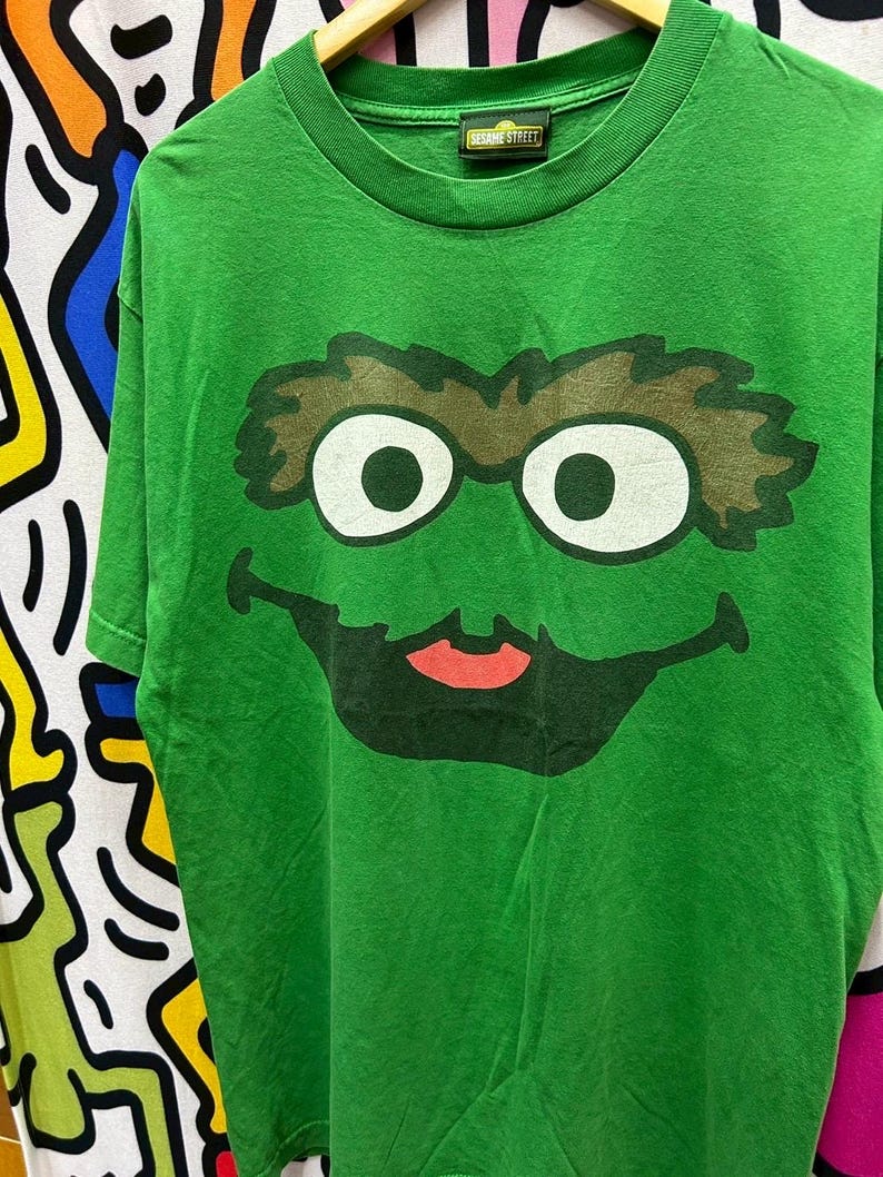 May include: Green Sesame Street t-shirt featuring a large graphic of Oscar the Grouch's face. The design includes white eyes, brown eyebrows, and a black mouth with a red tongue. The shirt has a Sesame Street tag.