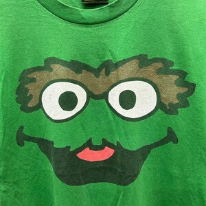 May include: Green t-shirt featuring a large graphic of Oscar the Grouch's face from Sesame Street. The design includes white eyes, brown eyebrows, a black mouth, and a red tongue. The Sesame Street label is visible at the neckline.