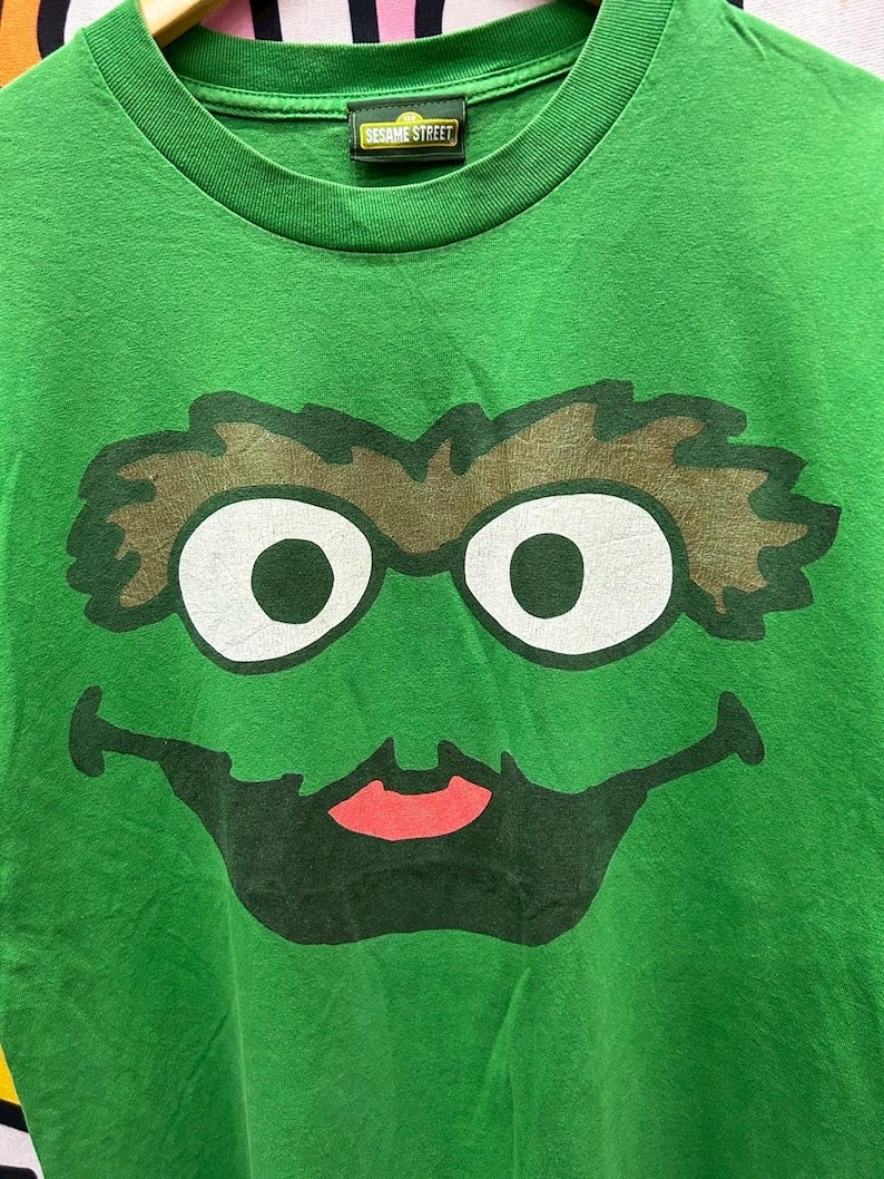 May include: Green t-shirt featuring a large graphic of Oscar the Grouch's face from Sesame Street. The design includes white eyes, brown eyebrows, a black mouth, and a red tongue. The Sesame Street label is visible at the neckline.