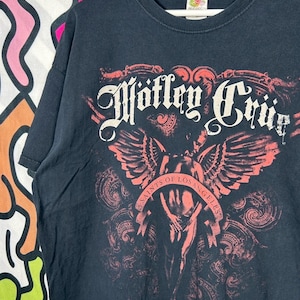以下が含まれることがあります： 赤と白のグラフィックプリントが施された黒の半袖Tシャツ。デザインには、スタイリッシュなフォントで書かれた「Mötley Crüe」の文字と、その下に「Saints of Los Angeles」と書かれたバナーを持つ翼のある人物のイラストが描かれています。