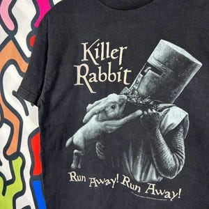 May include: A black short-sleeved t-shirt featuring a graphic of a knight in a helmet holding a rabbit. The text "Killer Rabbit" appears above the graphic, and the text "Run Away! Run Away!" is printed below the graphic in a light-coloured font.