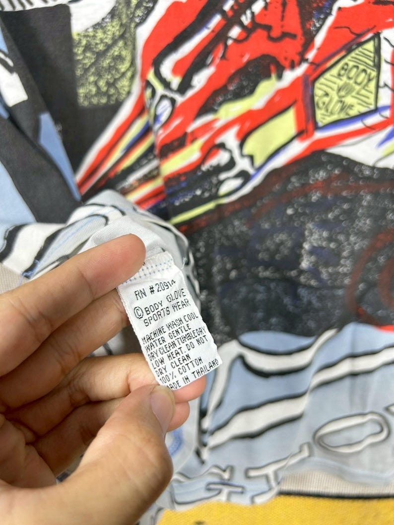 Puede incluir: Primer plano de una camiseta gr&aacute;fica colorida con la etiqueta "BODY GLOVE SPORTS WEAR". La camiseta presenta un dise&ntilde;o vibrante con elementos rojos, amarillos, azules y negros. La etiqueta tambi&eacute;n incluye instrucciones de cuidado.