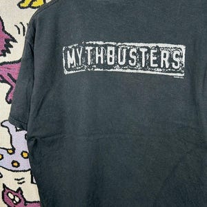 Puede incluir: Camiseta negra con el texto blanco "MYTHBUSTERS" en un rectángulo en la espalda. La camiseta es de material suave y tiene cuello redondo. El texto está ligeramente desgastado.