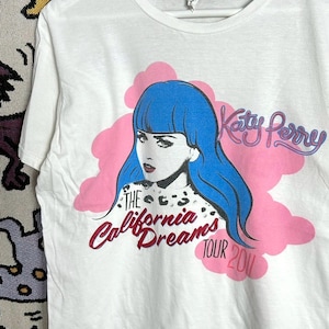 Könnte beinhalten: Weißes T-Shirt mit einer Grafik einer Frau mit blauen Haaren, vor einem rosa Wolken-Design. Der Text "Katy Perry" und "The California Dreams Tour 2011" sind ebenfalls auf dem T-Shirt aufgedruckt.