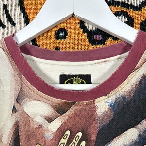 May include: Close-up of a crew neck t-shirt with a unique abstract design in shades of brown, beige, and blue. The neckline is trimmed with a burgundy band. A small black tag with yellow text is visible near the neckline.