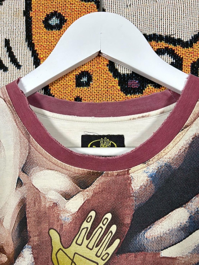 May include: Close-up of a crew neck t-shirt with a unique abstract design in shades of brown, beige, and blue. The neckline is trimmed with a burgundy band. A small black tag with yellow text is visible near the neckline.