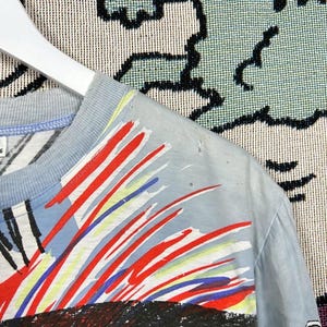 Puede incluir: Camiseta azul claro con un dise&ntilde;o abstracto colorido. El dise&ntilde;o presenta pinceladas rojas, blancas y azules sobre un gr&aacute;fico negro y rojo. La camiseta est&aacute; colgada de una percha blanca.