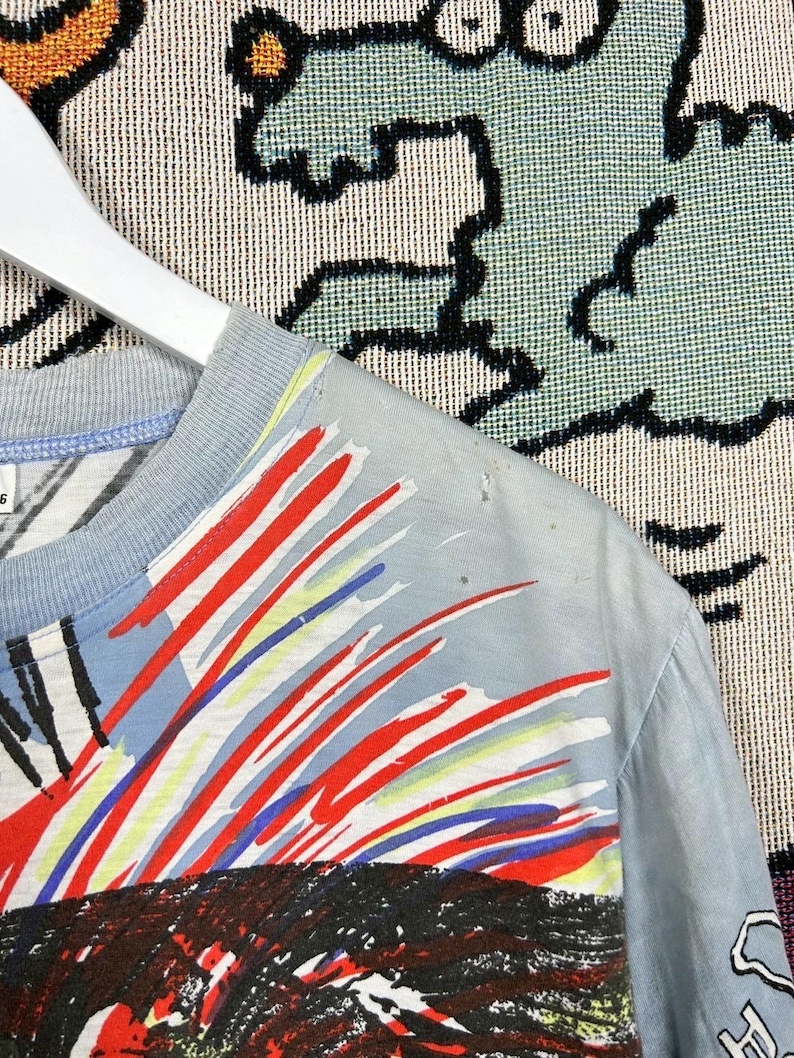 Puede incluir: Camiseta azul claro con un dise&ntilde;o abstracto colorido. El dise&ntilde;o presenta pinceladas rojas, blancas y azules sobre un gr&aacute;fico negro y rojo. La camiseta est&aacute; colgada de una percha blanca.
