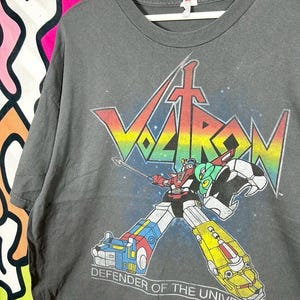 May include: A charcoal gray short-sleeve t-shirt featuring a graphic of the Voltron robot. The design includes the rainbow-colored Voltron logo, the robot holding a sword, and the text "DEFENDER OF THE UNIVERSE" printed in white at the bottom.