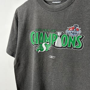 May include: A gray t-shirt with a green and white graphic that says "2007 Grey Cup Champions" with a hockey trophy and the text "Grey Cup" in a circle. The shirt is made by Reebok.