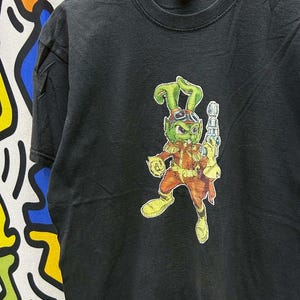 Könnte beinhalten: Schwarzes T-Shirt mit einer Cartoon-Figur mit grüner Haut, Brille und einer Strahlenkanone. Die Figur trägt ein rot-oranges Outfit mit gelben Stiefeln und Handschuhen. Das T-Shirt hängt an einem Holzkleiderbügel.