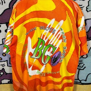 May include: A vintage Body Glove t-shirt with a vibrant orange and yellow tie-dye pattern. The shirt features the Body Glove logo in green and white, with the text "Body Glove" encircling the design. The shirt is hanging on a white hanger.