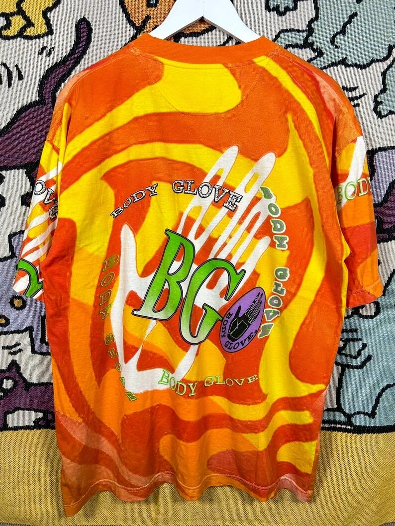 May include: A vintage Body Glove t-shirt with a vibrant orange and yellow tie-dye pattern. The shirt features the Body Glove logo in green and white, with the text "Body Glove" encircling the design. The shirt is hanging on a white hanger.