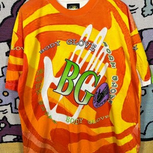 May include: Orange and yellow Body Glove t-shirt with a large white handprint graphic. The shirt features the text "BODY GLOVE" in a circular pattern around the handprint and "BG" in green letters. The shirt has a crew neck and short sleeves.
