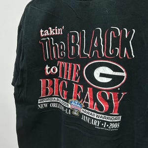 May include: Black t-shirt with a red and white graphic that says "takin' The BLACK to THE BIG EASY" with a Georgia Bulldogs logo and the text "GEORGIA BULLDOGS Allstate Sugar Bowl NEW ORLEANS LA HAWAII WARRIORS JANUARY 1-2008".