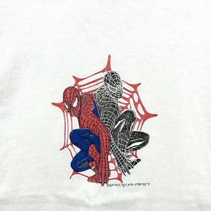 May include: White t-shirt with a graphic of two Spider-Man figures, one in red and blue, the other in black. They are set within a red and white spiderweb design, with the text "SPIDER-MAN" beneath the figures.