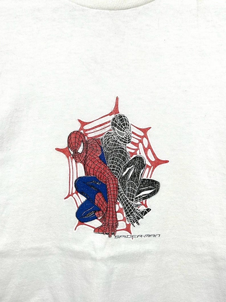 May include: White t-shirt with a graphic of two Spider-Man figures, one in red and blue, the other in black. They are set within a red and white spiderweb design, with the text "SPIDER-MAN" beneath the figures.