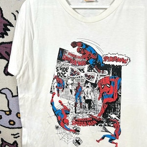 May include: White t-shirt featuring a colorful Spider-Man comic book graphic. The design includes multiple Spider-Man figures in action poses, comic book panels, and the text "SPIDER-MAN!" and "AMAZING SPIDER-MAN". The Marvel Comics logo is visible.