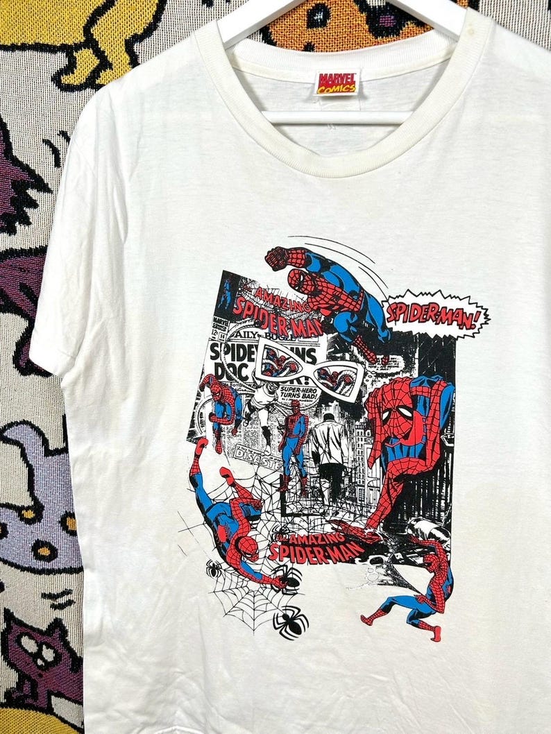 May include: White t-shirt featuring a colorful Spider-Man comic book graphic. The design includes multiple Spider-Man figures in action poses, comic book panels, and the text "SPIDER-MAN!" and "AMAZING SPIDER-MAN". The Marvel Comics logo is visible.