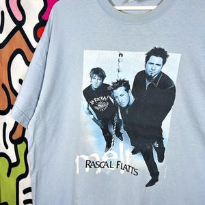 May include: A light blue short-sleeve t-shirt featuring a black and white graphic print of the country music band Rascal Flatts. The graphic includes the band members and the text "melt" and "RASCAL FLATTS" printed in black lettering.
