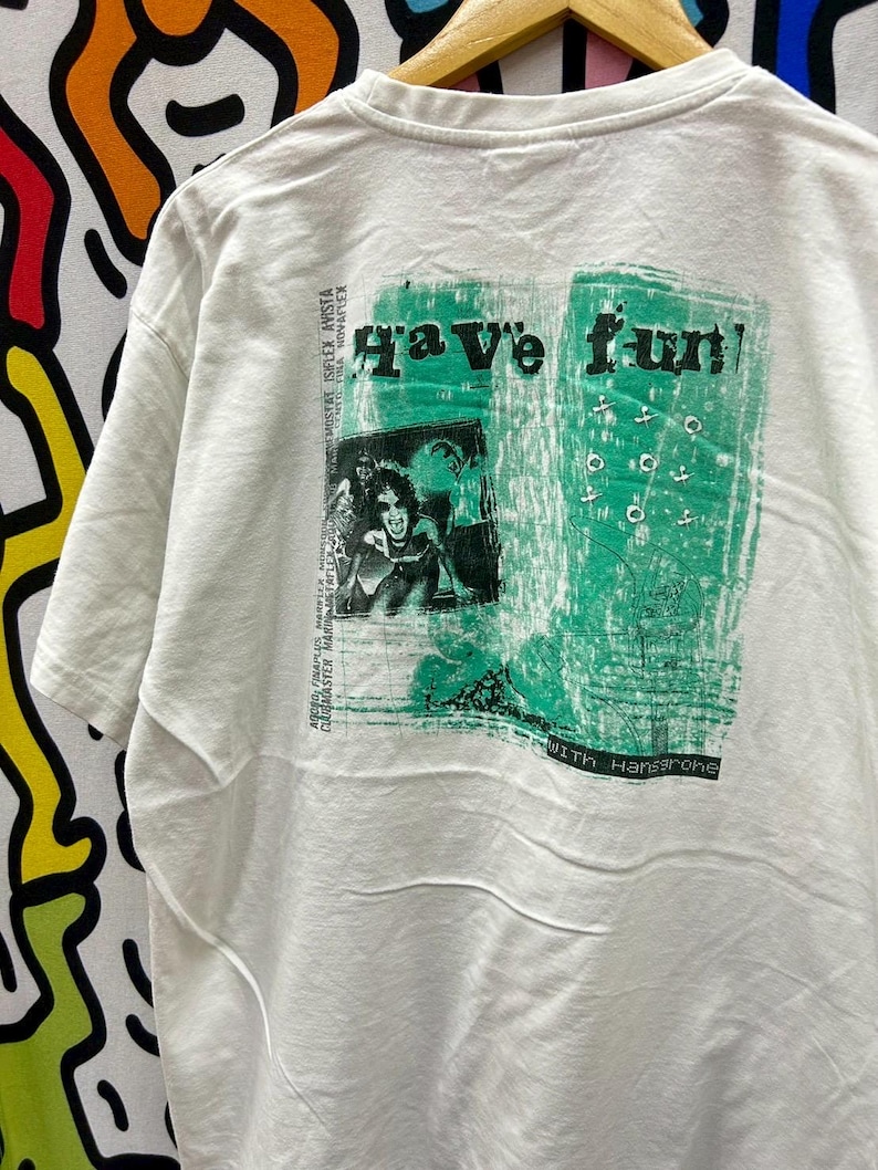 May include: White t-shirt with a green graphic print that says "Have fun!" with a picture of a person and the text "With Hansgrohe".