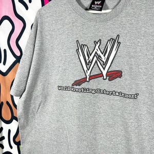 May include: A heather grey short-sleeve t-shirt featuring the white and black WWE logo with a red underline. Below the logo, the text World Wrestling Entertainment is printed in black. The shirt includes a black WWE brand neck label.