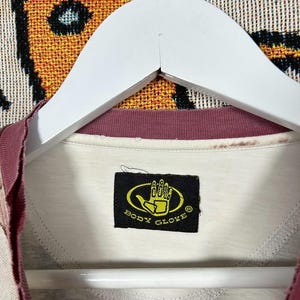 May include: Cream-colored Body Glove t-shirt with a burgundy collar and sleeve trim, hanging on a white hanger. The Body Glove logo is a yellow hand inside a black oval. The shirt is made of a soft material.