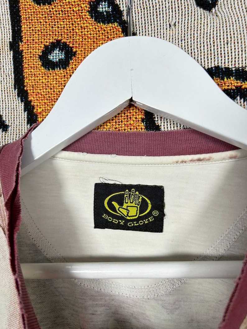 May include: Cream-colored Body Glove t-shirt with a burgundy collar and sleeve trim, hanging on a white hanger. The Body Glove logo is a yellow hand inside a black oval. The shirt is made of a soft material.