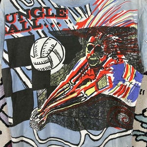 Puede incluir: Camiseta azul claro con un gr&aacute;fico colorido de un jugador de voleibol. La camiseta presenta las palabras "JUNGLE BALL" en rojo y blanco. El dise&ntilde;o incluye un voleibol y arte abstracto.
