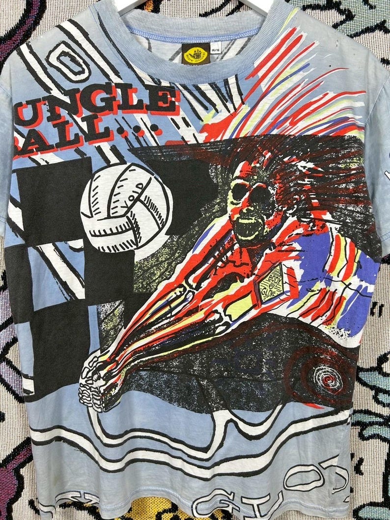 Puede incluir: Camiseta azul claro con un gr&aacute;fico colorido de un jugador de voleibol. La camiseta presenta las palabras "JUNGLE BALL" en rojo y blanco. El dise&ntilde;o incluye un voleibol y arte abstracto.